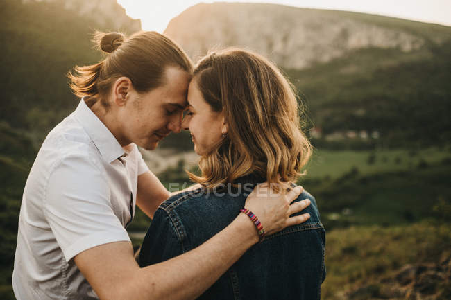 Linda pareja abrazando y endureciendo la frente mientras se sienta en la ladera rocosa en el fondo del hermoso valle y las montañas - foto de stock