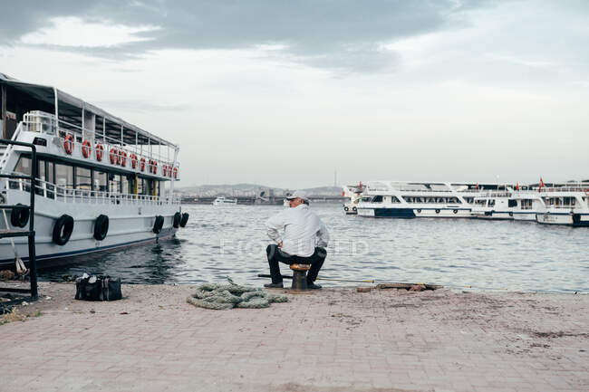 Vista trasera del hombre adulto sentado en el muelle cerca del agua y los barcos y la pesca en Estambul, Turquía - foto de stock