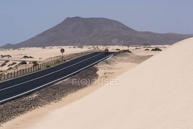 Larga carretera recta sobre llanura con dunas de arena con montañas, Islas Canarias - foto de stock