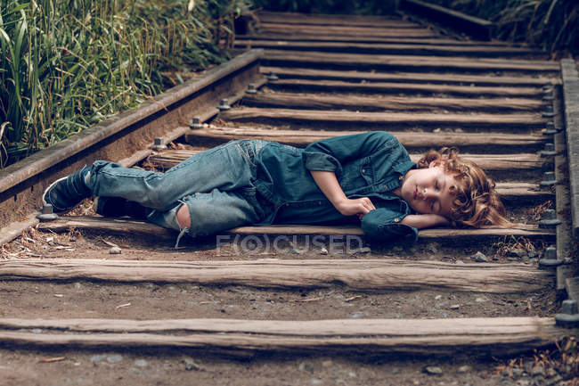Lonely adorable boy in denim sleeping on railway road lying on wooden beam in green grass — Stock Photo