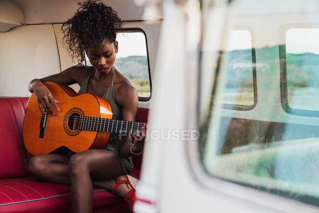 Hermosa mujer afroamericana sentada en el asiento trasero rojo de la furgoneta retro y tocando la guitarra acústica mientras viaja en la naturaleza - foto de stock