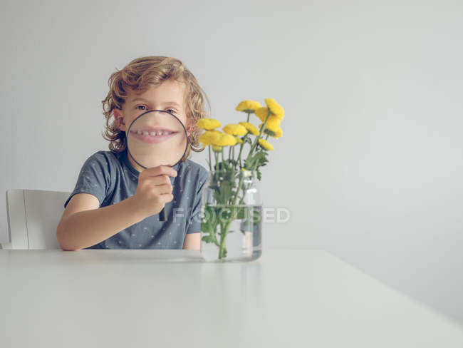 Frasco com dentes-de-leão frescos em pé na mesa perto do menino adorável se divertindo com lupa e olhando para a câmera — Fotografia de Stock