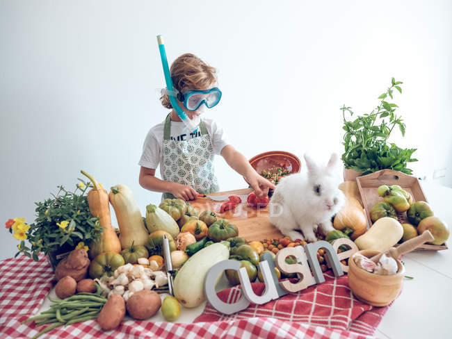Молодий хлопчик стоїть і готує овочі за столом з чарівним білим кроликом — стокове фото