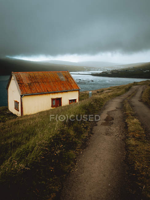 Pequeña casa gruñona con techo oxidado en la orilla del lago en las Islas Feroe en día nublado - foto de stock