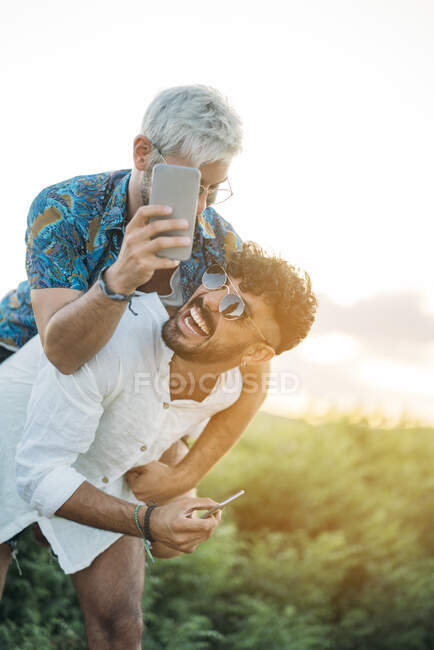 Handsome bearded guy giving piggyback ride to cheerful boyfriend and posing for selfie while spending time in nature together — Stock Photo
