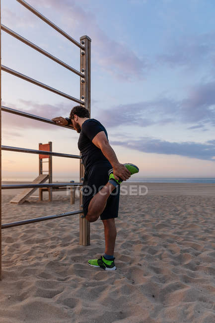 Chico musculoso en ropa deportiva haciendo ejercicio de calentamiento para las piernas mientras está de pie cerca de la escalera durante la puesta de sol en la playa - foto de stock