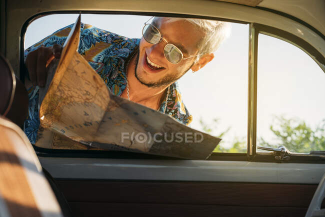 Joven guapo con mapa sonriendo y mirando dentro de coche vintage mientras viaja por el campo - foto de stock