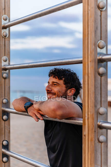 Lächelnder bärtiger Mann in Sportbekleidung lehnt auf Leiter und schaut weg, während er sich beim Outdoor-Training ausruht — Stockfoto