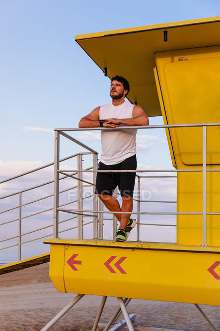 Homme barbu en vêtements de sport appuyé sur la rampe de la cabine de sauveteur sur la plage pendant l'entraînement en plein air au coucher du soleil — Photo de stock