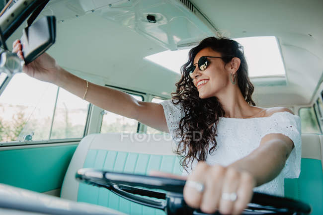 Sorrindo jovem mulher dirigindo carro retro e verificando retrovisor espelho — Fotografia de Stock