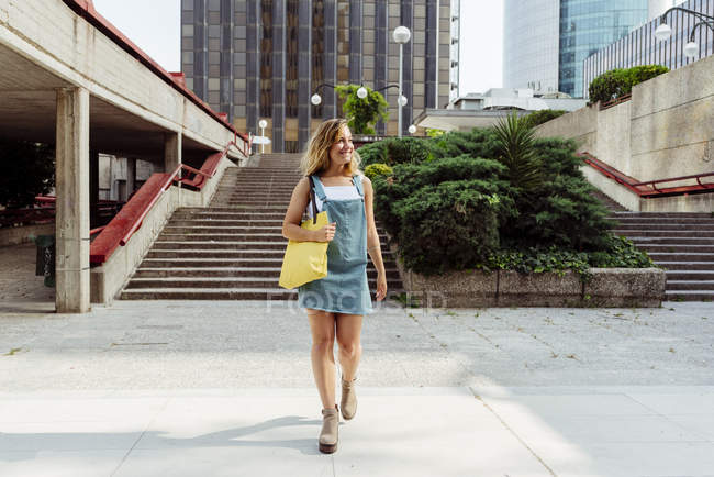 Cheerful young casual dressed woman with yellow handbag walking on city street and smiling — Stock Photo