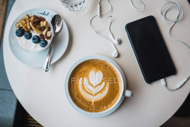 Зверху чашка кави з фруктовим десертом і смартфон, покладений поруч на столі — стокове фото
