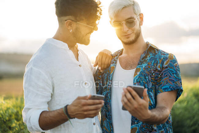 Zwei lächelnde Männer, die sich umarmen und Smartphones schmökern, während sie gemeinsam in der schönen Landschaft stehen — Stockfoto