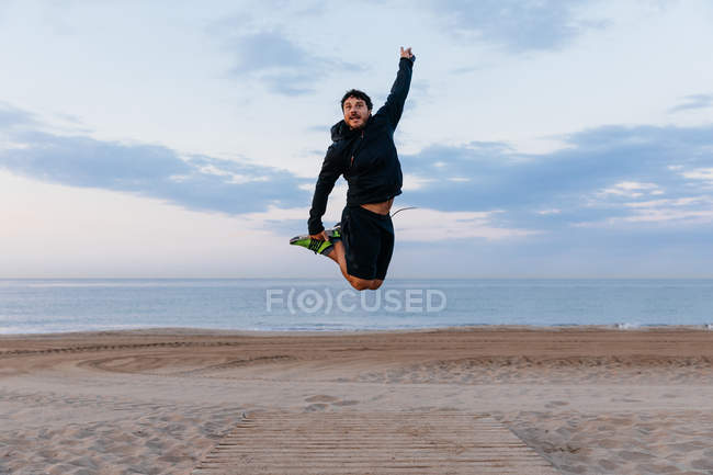 Homme barbu actif sautant sur la plage au coucher du soleil — Photo de stock