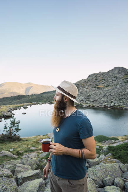 Junger Mann mit Hut steht mit Tasse am See und blickt auf die Aussicht — Stockfoto