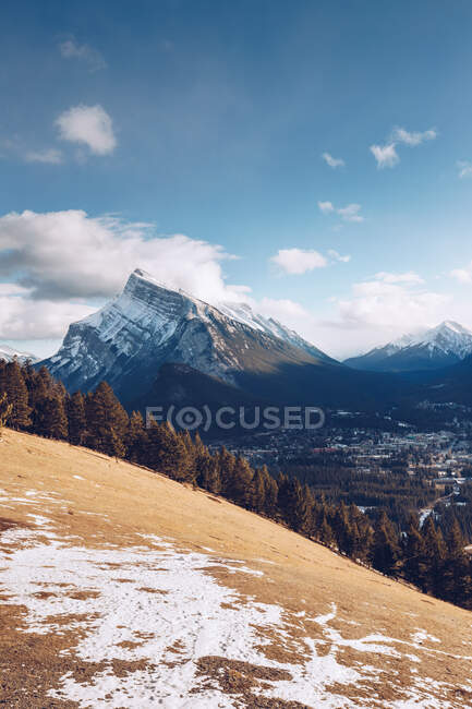 Hill with yellow frozen soil and thick forest on background with snowy mountains and blue sky with few clouds and valley with small city — Stock Photo