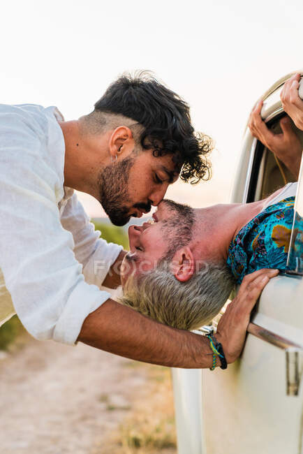 Side view of man sitting inside of car and leaning out of window kissing with boyfriend standing outside in summertime — Stock Photo