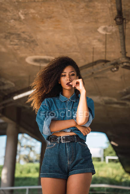 Mujer joven en ropa de mezclilla de pie debajo del puente y mirando a la cámara - foto de stock