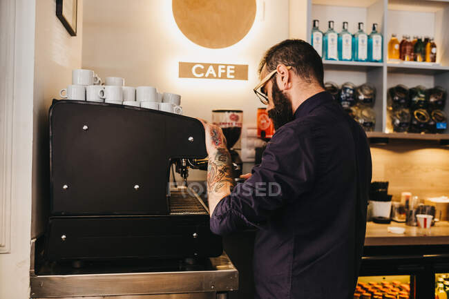 Side view of tattooed bearded man working as barista in coffee shop and brewing coffee with machine — Stock Photo