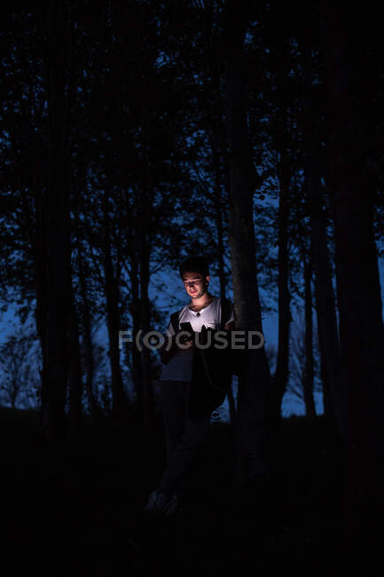 Guapo joven que se apoya en el tronco del árbol y el teléfono inteligente de navegación mientras pasa tiempo en el bosque por la noche - foto de stock