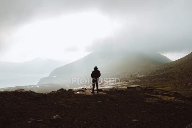 Homem caminhante irreconhecível em pé na costa sob nuvens e olhando para o oceano nas Ilhas Feroé — Fotografia de Stock