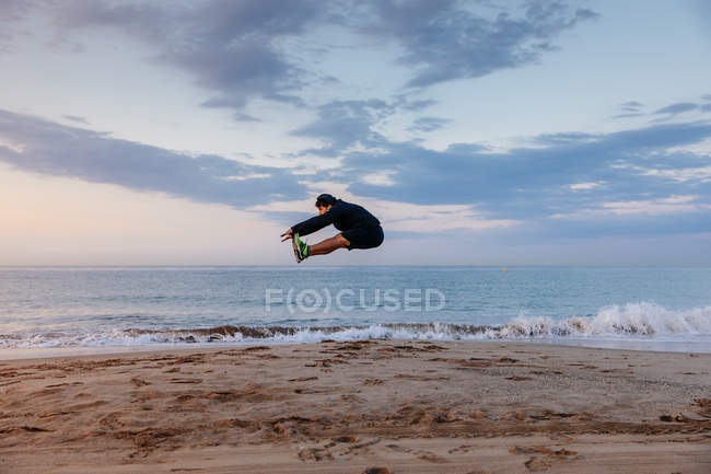 Active man in sportswear jumping high during outdoor training on sandy beach at sunset — Stock Photo