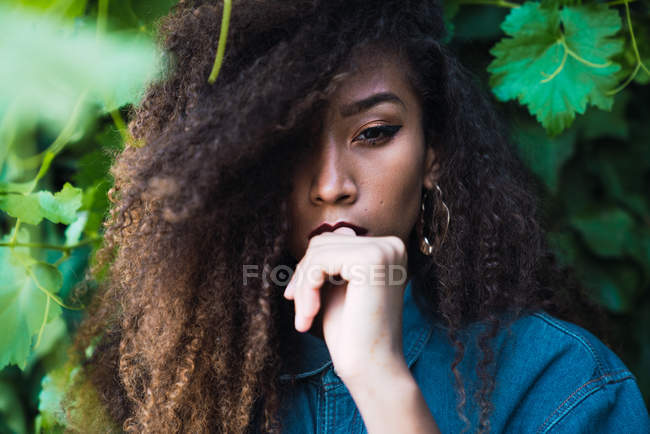 Sensual mujer afroamericana de pie contra el arbusto verde y mirando a la cámara - foto de stock