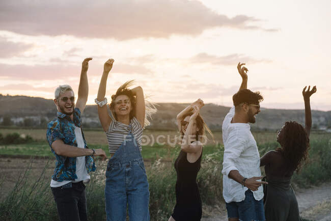Группа молодых людей в повседневной одежде смеются и танцуют, веселясь в красивой сельской местности вместе — стоковое фото