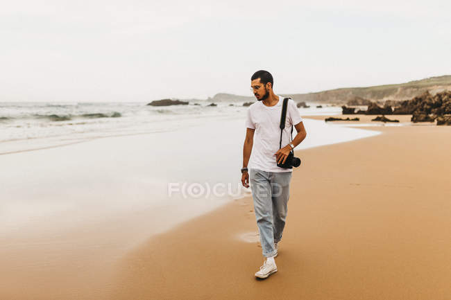 Jovem atraente com câmera fotográfica andando na praia de areia e olhando para a água do mar enquanto viaja na natureza — Fotografia de Stock