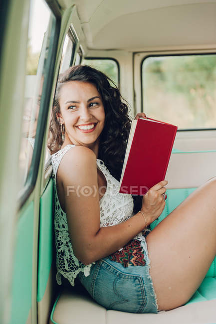 Sonriente joven sentada dentro de la caravana y leyendo con libro - foto de stock