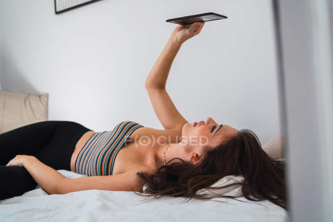 Молодая брюнетка в полосатом топе и черных колготках лежит на кровати в комнате и использует смартфон — стоковое фото