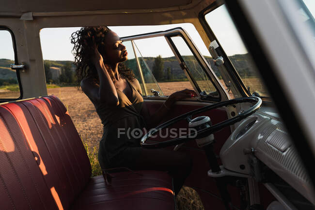 Прекрасная афроамериканка улыбается и смотрит в окно винтажного автомобиля, проводя время на природе в солнечный день — стоковое фото