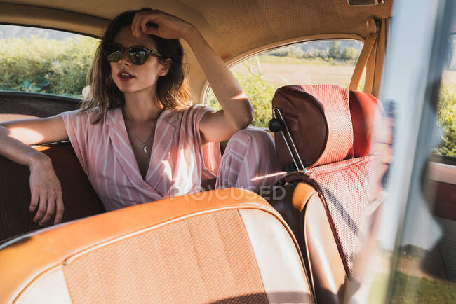 Уверенная женщина в розовых очках и солнцезащитных очках, сидящая на заднем сиденье в ретро-машине и отводящая взгляд от солнца — стоковое фото