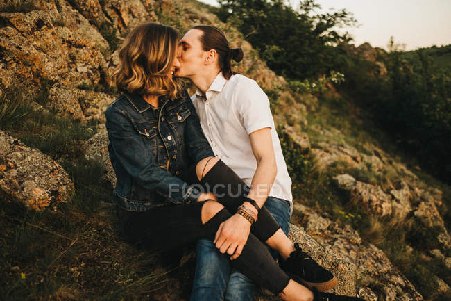 Jeune homme embrassant petite amie dans la joue tout en étant assis ensemble sur la pente de la montagne rocheuse — Photo de stock