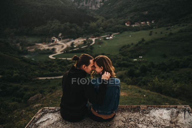 Cute couple hugging and toughing foreheads while sitting on rocky slope on background of beautiful valley and mountains — Stock Photo