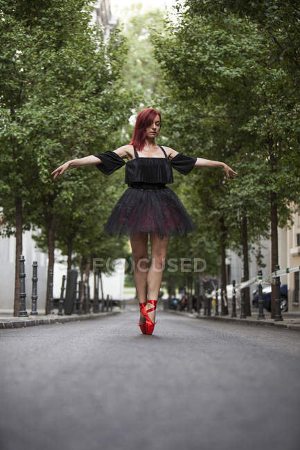 Ballerine à tête rouge avec tutu noir et pointes de ballet rouge dansant dans la rue avec des arbres en arrière-plan . — Photo de stock