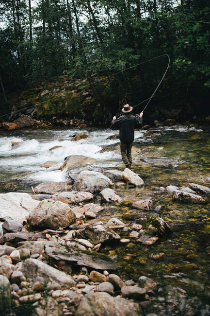 Pescador sosteniendo varilla giratoria y de pie sobre rocas en el flujo del río - foto de stock