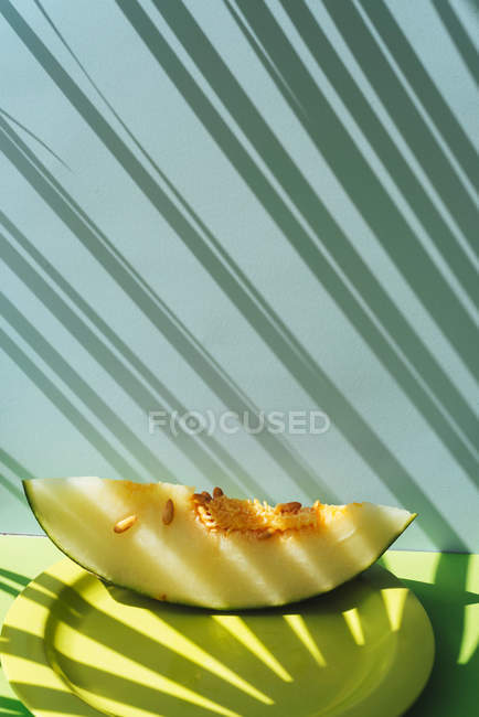 Fatia de melão fresco na placa no fundo azul e verde com sombras de folhas de palma — Fotografia de Stock