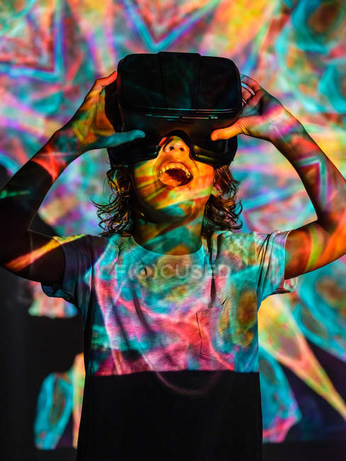 Garoto bonito vestindo óculos VR e explorando a realidade virtual com expressão facial animada enquanto está sob projeção colorida — Fotografia de Stock
