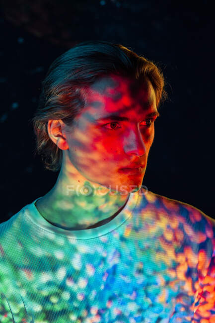 Jeune homme androgyne illuminé avec des sports colorés de lumière vive regardant loin tout en se tenant sur fond noir — Photo de stock