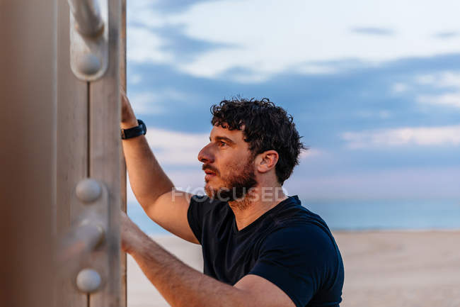 Бородатый мужчина в спортивной одежде опирается на лестницу и смотрит в сторону во время отдыха на открытом воздухе на закате — стоковое фото