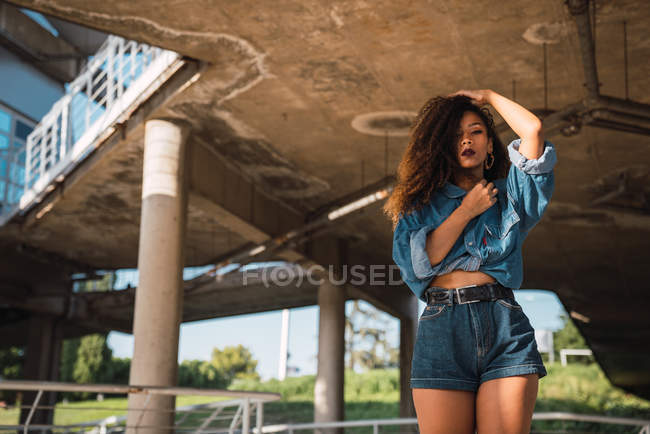 Молодая афроамериканка в джинсовой рубашке и шортах, стоящая под мостом с рукой в волосах и смотрящая в камеру — стоковое фото
