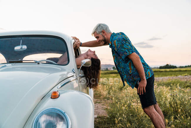 Side view of woman sitting inside of car leaning head out and looking at man standing above and smiling happily in summertime — Stock Photo