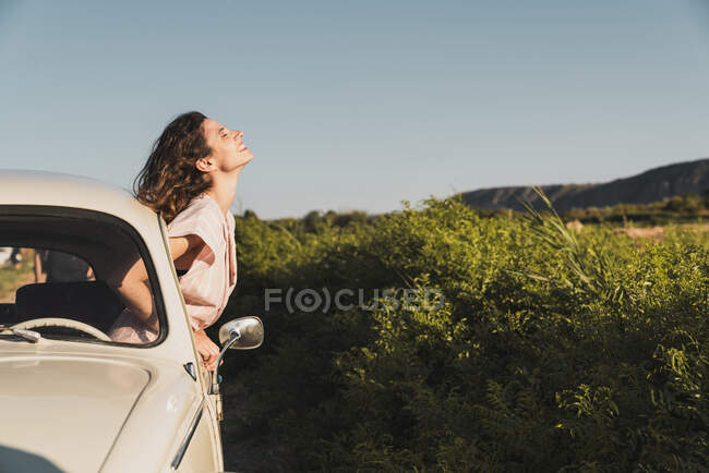 Mujer joven feliz en gafas de sol elegantes mirando por la ventana del coche disfrutando de la luz del sol de verano contra árboles verdes y el cielo azul - foto de stock