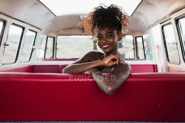 Приваблива афро-американська жінка сидить на зручному задньому сидінні вінтажного фургона і озирається геть під час подорожі в сільській місцевості. — стокове фото