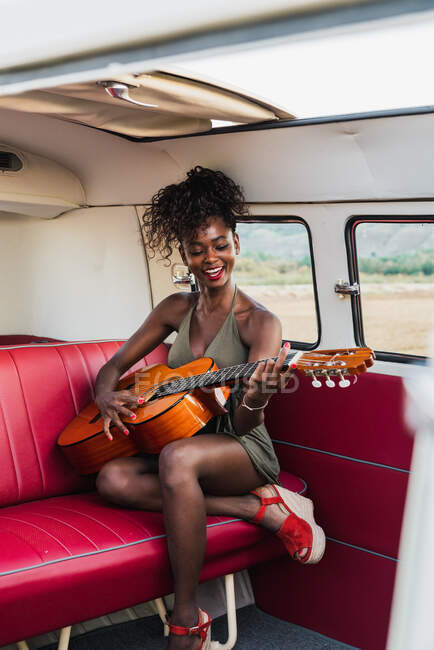 Beautiful African-American woman sitting on red back seat of retro van and playing acoustic guitar while traveling in nature — Stock Photo