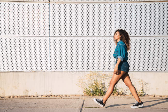 Young funky African American woman with dark hair in denim clothes and sneakers walking on street near wall at sunny day — Stock Photo