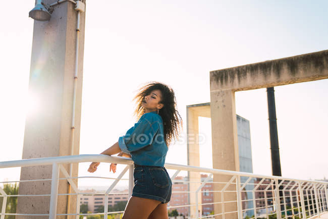 Молода чуттєва жінка в джинсовому одязі, що стоїть на балконі біля перил на сонячному світлі — стокове фото