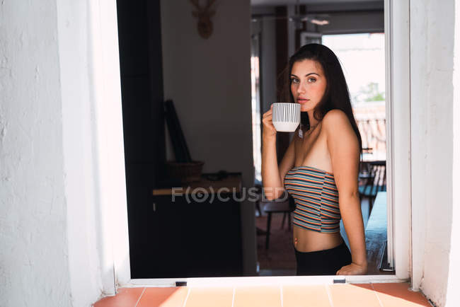 Young brunette woman in crop top standing near window and drinking coffee on background of room — Stock Photo