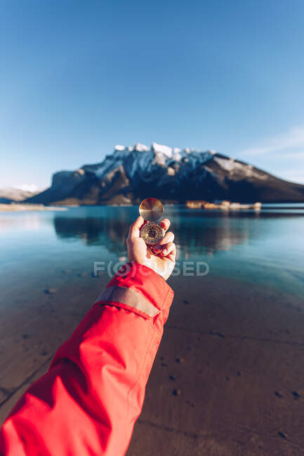 Humano vestido con chaqueta roja sosteniendo brújula dorada en un día soleado sobre un fondo borroso de montañas canadienses - foto de stock